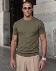 Man wearing olive green knit t-shirt by a stone wall, Morengar summer menswear